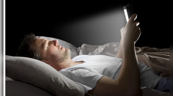 Electronics emitting blue light on a nightstand next to a bed.